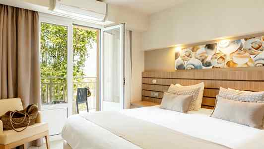 Hotellihuone, jossa suuri sänky, istuinkaluste ja avoin parvekkeen ovi näkymällä vehreyteen. (Tekoälyn luoma teksti)