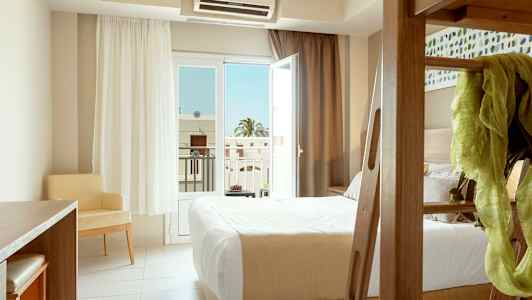 Hotellihuone, jossa on sänky, kirjoituspöytä ja tuoli, sekä avoin lasi-ikkuna parvekkeelle. (Tekoälyn luoma teksti)