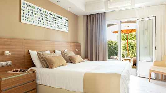 Valoisa hotellihuone, jossa on iso sänky, seinätaidetta ja parvekkeen ovet näkymällä puutarhaan, jossa on oranssi aurinkovarjo. (Tekoälyn luoma teksti)