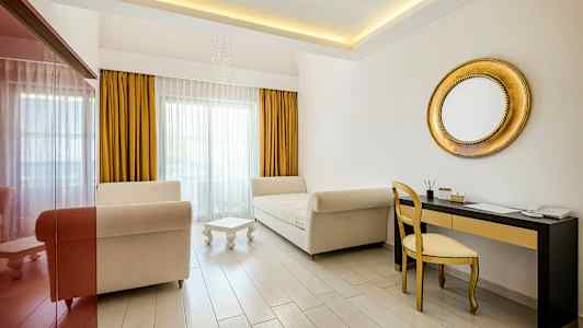 Kuva valoisasta huoneesta, jossa on kaksi sohvaa, pieni pöytä, kullanvärinen peili ja kirjoituspöytä tuolilla. (Tekoälyn luoma teksti)