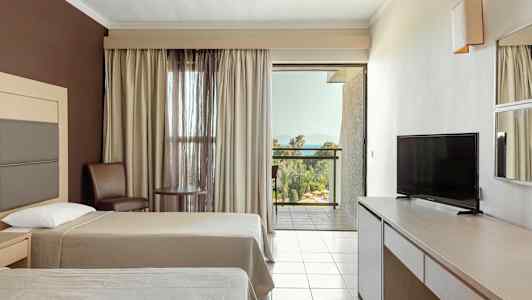 Hotellihuone, jossa on kaksi sänkyä, TV, pöytä ja tuoleja sekä parveke näkymällä vehreään maisemaan ja merelle. (Tekoälyn luoma teksti)