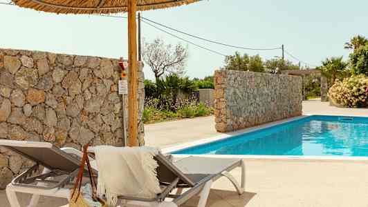 Kaksi aurinkotuolia, joissa on pyyhe ja olkilaukku, uima-altaan vieressä. Taustalla on myös kivimuuri ja palmuja. (Tekoälyn luoma teksti)