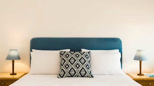 Sinisellä päätyllä varustettu sänky, jossa on valkoiset tyynyt ja koristeellinen tyyny keskellä. Molemmilla puolilla yöpöydät, joissa lamput. (Tekoälyn luoma teksti)