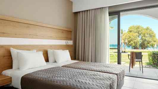 Kaksi vuodetta, joissa valkoisia tyynyjä ja harmaat peitteet, lasiovien vieressä, jotka johtavat puutarhaan merinäköalalla. (Tekoälyn luoma teksti)