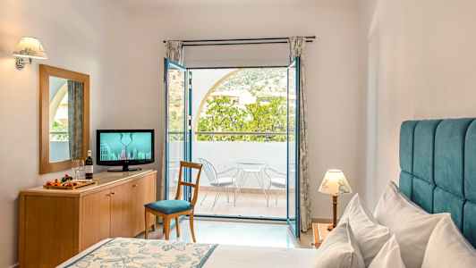 Hotellihuone, jossa on sänky, lipasto, jossa televisio, ja parveke, jolla on tuolit ja pöytä. (Tekoälyn luoma teksti)