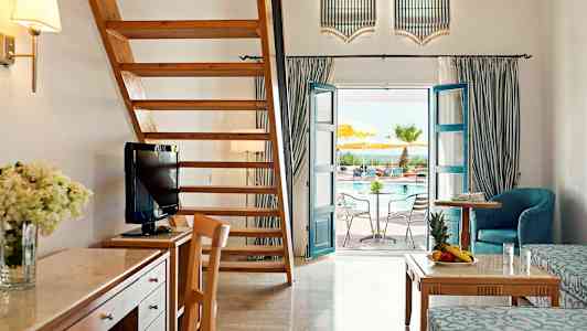 Tilava hotellihuone, jossa on puisia huonekaluja, puuportaat ja ovi, joka johtaa uima-altaalle. (Tekoälyn luoma teksti)