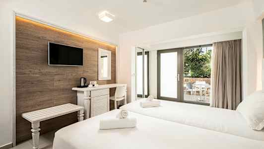 Hotellihuone, jossa on kaksi vuodetta, kirjoituspöytä, televisio ja lasiovien kautta näkyvä ulkoterassi, jossa on pöytä ja tuoleja. (Tekoälyn luoma teksti)