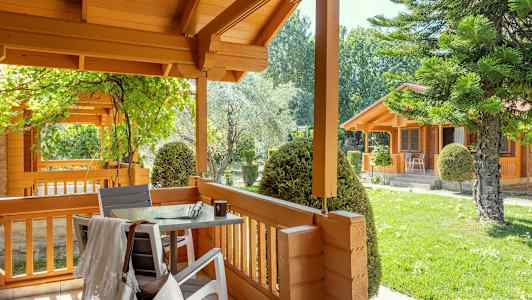 Puisella verannalla on pöytä ja tuolit, ja sitä ympäröivät vehreä ympäristö ja puiset talot. (Tekoälyn luoma teksti)