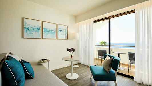 Vaalea olohuone, jossa harmaa sohva, sinisiä koristeellisia tyynyjä ja pöytä sekä näkymä parvekkeelle ja merelle. (Tekoälyn luoma teksti)