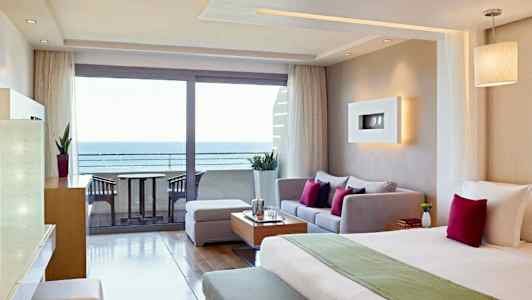 Hotellihuone, jossa on moderni sisustus, sisältäen sängyn, oleskelutilan ja merinäköalan lasiovien kautta. (Tekoälyn luoma teksti)