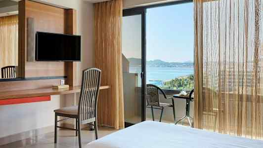 Hotellihuone, jossa on kirjoituspöytä, tuoli ja TV. Ikkuna ja parveke merinäköalalla. (Tekoälyn luoma teksti)