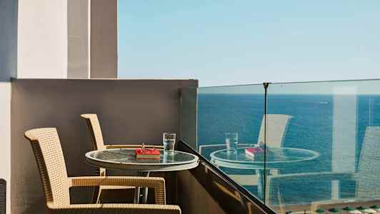 Näkymä parvekkeelta, jossa on kaksi tuolia ja pöytä, johon on asetettu vesilasi ja kirja meren edessä. (Tekoälyn luoma teksti)