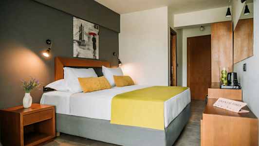 Makuuone, jossa on parivuode, valkoiset ja keltaiset vuodevaatteet, yhteensopivat tyynyt, yöpöytä maljakolla, jossa kukkia, sekä seinällä sängyn yläpuolella taulu. (Tekoälyn luoma teksti)