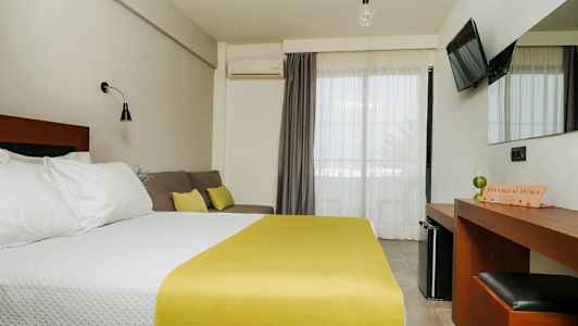 Hotellihuone, jossa on parivuode, valkoiset vuodevaatteet ja keltainen päiväpeite, sohva tyynyineen, työpöytä peileineen ja seinälle kiinnitetty TV. Parvekkeen ovet vaaleilla verhoilla. (Tekoälyn luoma teksti)