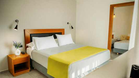 Makuuhuone, jossa on parivuode, kaksi valkoista tyynyä ja keltainen peitto. Yöpöydällä on valkoinen maljakko ja kasvi. (Tekoälyn luoma teksti)