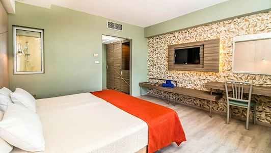 Hotellihuone, jossa on parivuode, valkoiset ja oranssit vuodevaatteet, geometristen yksityiskohtien seinä sekä televisio ja pöytä. (Tekoälyn luoma teksti)