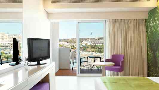 Hotellihuone, josta on näkymä merelle. Kalusteina televisio, työpöytä, violetti tuoli ja suuri sänky. (Tekoälyn luoma teksti)