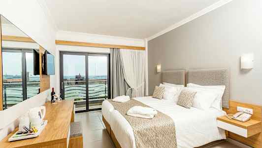 Hotellihuone, jossa iso sänky, vaaleat kalusteet ja parveke näkymällä. (Tekoälyn luoma teksti)