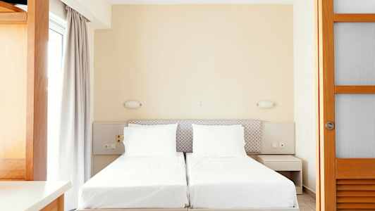 Kaksi yksittäistä sänkyä valkoisilla vuodevaatteilla ja neutraaleja huonekaluja. (Tekoälyn luoma teksti)