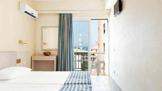 Hotellihuone, jossa kaksi sänkyä, sinikeltaiset päiväpeitot, peili ja parvekeovi, josta avautuu näkymä. (Tekoälyn luoma teksti)