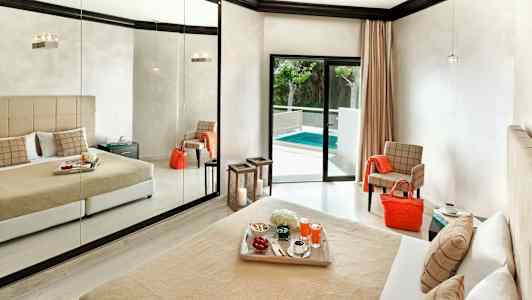 Hotellihuone, jossa suuri peiliseinä, beige vuodepeite ja tyynyt sekä parvekkeen ovi, joka johtaa uima-altaalle. (Tekoälyn luoma teksti)