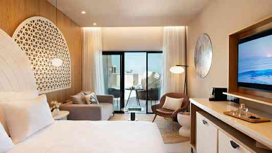 Moderni hotellihuone, jossa on vaaleat kalusteet ja koristeet, sisältäen sängyn, sohvan, tuolin ja television. Huoneessa on suuret lasiovet, jotka johtavat parvekkeelle. (Tekoälyn luoma teksti)