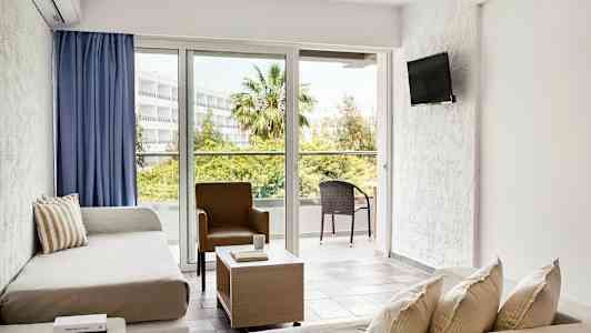 Valoisa hotelli huone, jossa on oleskelutila, sohvat, tuoli, sohvapöytä ja suuret ikkunat, jotka avautuvat parvekkeelle ulkokalusteilla vehreässä ympäristössä. (Tekoälyn luoma teksti)