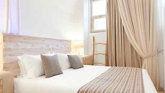 Vaaleasävyinen makuuhuone, jossa suuri sänky, tyynyjä ja yksityiskohtainen puusisustus. (Tekoälyn luoma teksti)