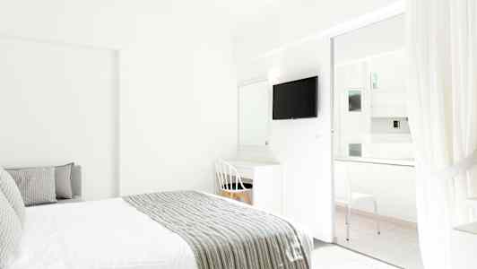 Valoisan hotellihuoneen sisustus, jossa on sänky ja kirjoituspöytä valkoisen tuolin vieressä. TV on kiinnitetty seinään, ja lasiovet vievät parvekkeelle. (Tekoälyn luoma teksti)