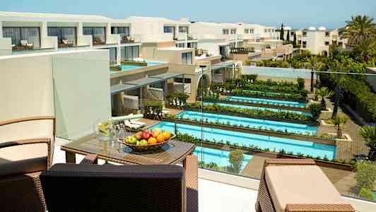 Hotellialue, jossa useita uima-altaita ja viheralueita, sekä terassi kalusteineen ja hedelmin. Näkymä moderneihin rakennuksiin. (Tekoälyn luoma teksti)