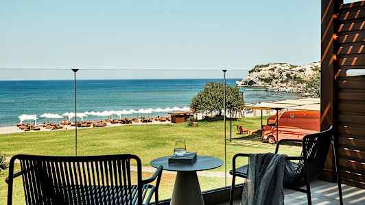 Näkymä terassilta vihreän nurmikon yli rannalle, jossa on aurinkotuoleja ja aurinkovarjoja, sekä meri ja kallioita taustalla. (Tekoälyn luoma teksti)