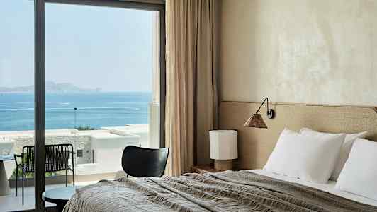 Hotellihuone, jossa on sänky, pöytä ja tuoli, merinäköala suurten ikkunoiden läpi. (Tekoälyn luoma teksti)