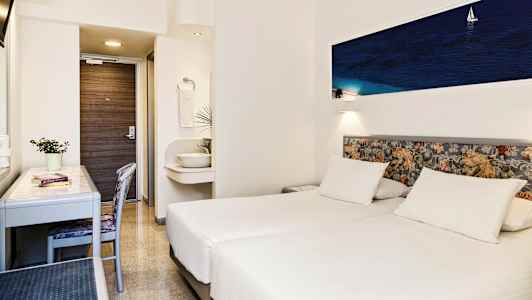 Hotellihuone, jossa on kaksi valkoista yhden hengen vuodetta, kirjoituspöytä, tuoli ja pieni pesuallas kylpyhuoneen ulkopuolella. (Tekoälyn luoma teksti)