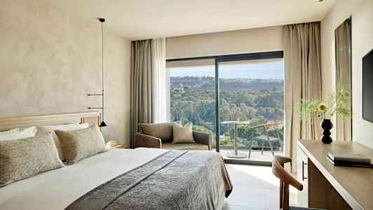 Moderni hotellihuone, jossa on suuri sänky, oleskelutila ja parveke, josta avautuu näkymä vehreään maisemaan. (Tekoälyn luoma teksti)