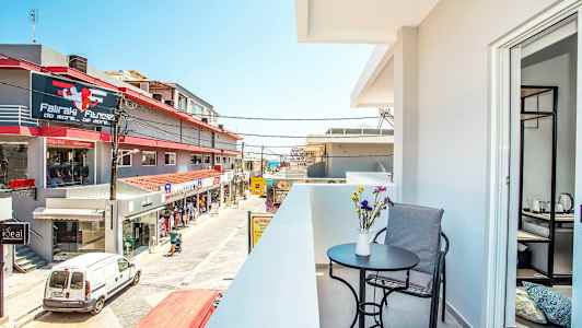 Parveke, jolla on pöytä, tuoli ja kukkia etualalla, sekä näkymä kadulle, jossa on liikkeitä ja rakennuksia. (Tekoälyn luoma teksti)