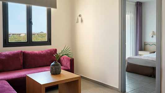 Huone, jossa on violetti kulmasohva, pöytä maljakolla ja viereinen makuuhuone näkyvissä oviaukosta. (Tekoälyn luoma teksti)