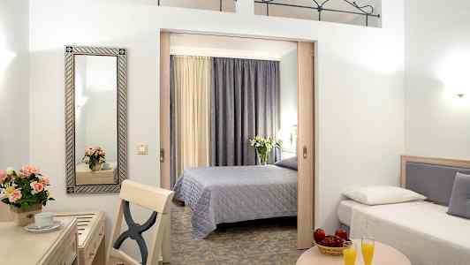 Hotellihuone, jossa on kaksi sänkyä, pöytä, jonka päällä on kaksi lasillista appelsiinimehua, sekä kirjoituspöytä, jossa on peili ja kukkia. (Tekoälyn luoma teksti)