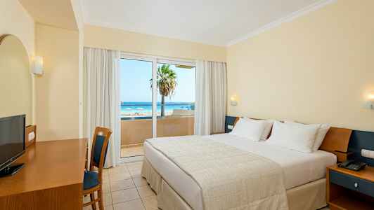 Hotellihuone, jossa on parivuode, kirjoituspöytä, tuoli ja näkymä rannalle lasioven läpi. (Tekoälyn luoma teksti)