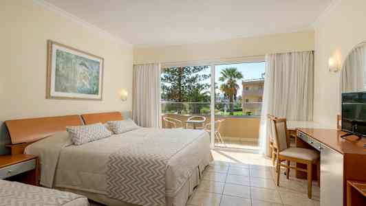 Hotellihuone, jossa on pedattu sänky, kirjoituspöytä ja tuoleja lasioven vieressä, joka johtaa parvekkeelle. (Tekoälyn luoma teksti)