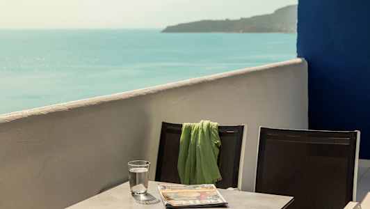 Kaksi tuolia, joiden yhteen on asetettu vihreä huopa, pöydän ääressä, jossa on vesilasi ja lehti, ja näkymä merelle. (Tekoälyn luoma teksti)