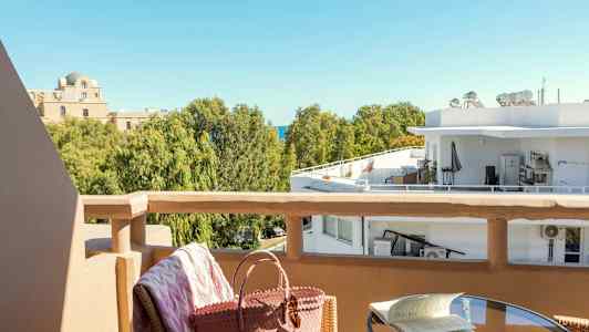 Näköala parvekkeelta puille ja rakennuksille. Etualalla pöytä, jossa kirja, ja tuoli, jossa on laukku. (Tekoälyn luoma teksti)