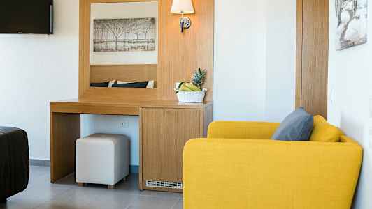 Hotellihuone, jossa keltainen nojatuoli, kirjoituspöytä ja hedelmäkulho. (Tekoälyn luoma teksti)