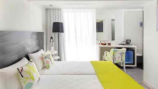 Valoisa hotellihuone, jossa on kaksi erillistä vuodetta, koristeelliset tyynyt ja limenvihreä peitto sekä istuinpaikka ja kirjoituspöytä peilillä. (Tekoälyn luoma teksti)