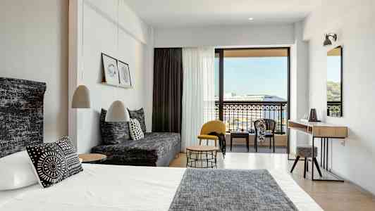 Moderni hotellihuone, jossa sänky, sohva ja istuinryhmä sekä näkymä parvekkeelle ja ympäristöön. (Tekoälyn luoma teksti)