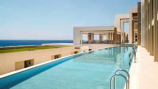 Kuva uima-altaasta modernin, vaalean rakennuskompleksin yhteydessä. Taustalla näkyy meri. (Tekoälyn luoma teksti)