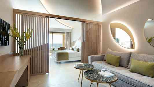 Hotellihuone, jossa istuinryhmä, pöytiä ja sänky taustalla. (Tekoälyn luoma teksti)