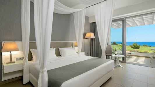 Hotellihuone, jossa on leveä sänky, valkoiset verhot ja näkymä merelle suurien ikkunoiden läpi. (Tekoälyn luoma teksti)