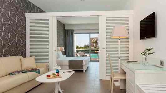 Hotellihuone, jossa sohva, pöytä koriste-esineillä, työpöytä ja tuoli. Taustalla oleva vuode johtaa terassille, jolta on näkymä altaalle. (Tekoälyn luoma teksti)