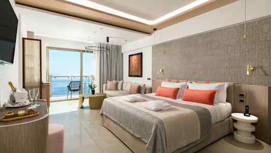 Moderni hotellihuone, jossa on pedattu sänky oransseilla tyynyillä, istuinalue ja parveke merinäkymin. (Tekoälyn luoma teksti)