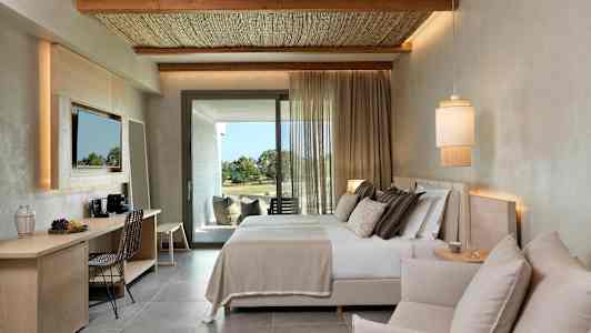 Moderni hotellihuone, jossa on parivuode, istuinryhmä ja kirjoituspöytä. Tekstiilit päästävät päivänvalon parvekkeen ovelta. (Tekoälyn luoma teksti)